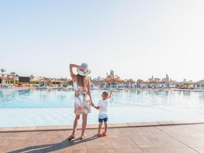 Frau mit zwei Kindern am Beckenrand eines hellen Hotels bei sonnigem Himmel.