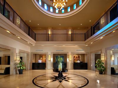 Helle Hotellobby mit Kronleuchter, runder Decke und Empfangstresen mit Pflanzen.
