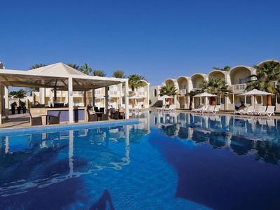 Ein großer Pool mit klarem Wasser vor einem weißen Hotel mit Bögen und Sonnenliegen am Rand.
