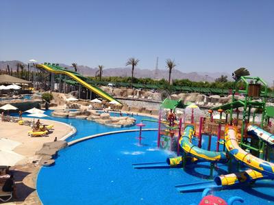 Wasserpark mit Rutschen, Pool und Sonnenschirmen vor bergiger Landschaft bei klarem Himmel.