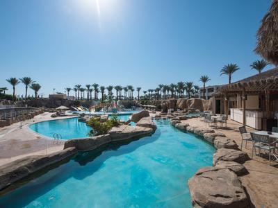 Poolbereich mit klarem Wasser, Felsen und Palmen unter strahlend blauem Himmel.