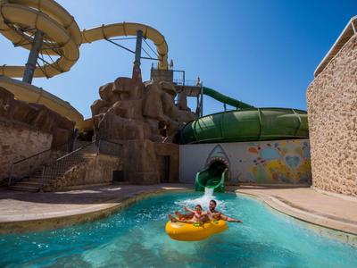 Kinder rutschen auf einer gelben Schwimmreifenrutsche in einem hellen Pool mit blauem Himmel.