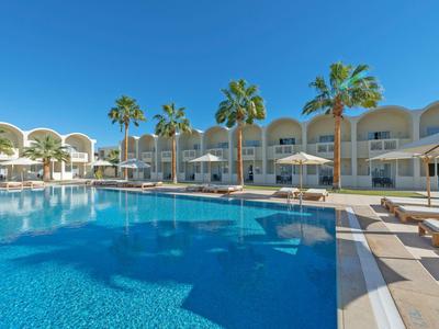 Großer Hotelpool mit Sonnenschirmen und Palmen vor modernen, weißen Gebäuden unter klarem Himmel.