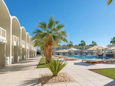 Moderner Hotelpoolbereich mit Liegestühlen, Sonnenschirmen und Palmen unter blauem Himmel.