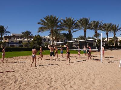 Personen spielen Volleyball auf Sandplatz vor Hotel mit Palmen unter blauem Himmel.