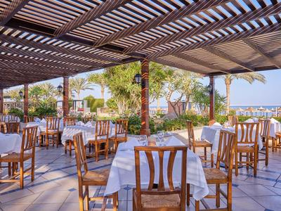 Freiluftrestaurant mit Holztischen, weißen Tischdecken und Blick auf Palmen und Meer.
