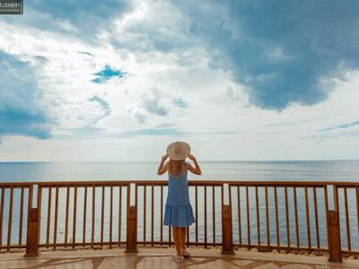 Frau in blauem Kleid mit Hut blickt auf das weite Meer von einer Holzterrasse aus.
