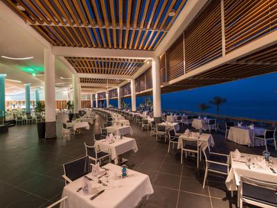 Modernes, überdachtes Restaurant mit weißen Tischen und Blick auf das Meer bei Dämmerung.