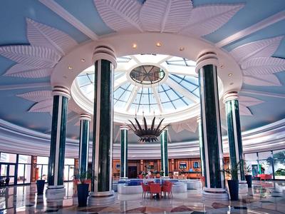 Luminosa hall d'hotel con alti pilastri, soffitto in vetro circolare e sedute moderne al centro.