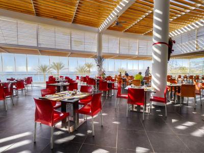 Helles, modernes Restaurant mit roten Stühlen und großen Fenstern, Sonnenlicht fällt ein.