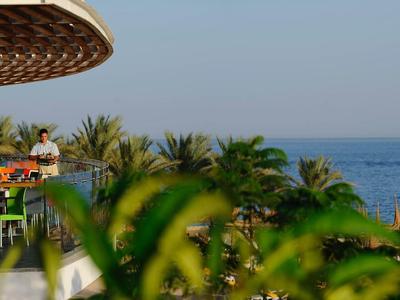 Vista di una terrazza con sedie e tavoli, circondata da palme e con il mare sullo sfondo.