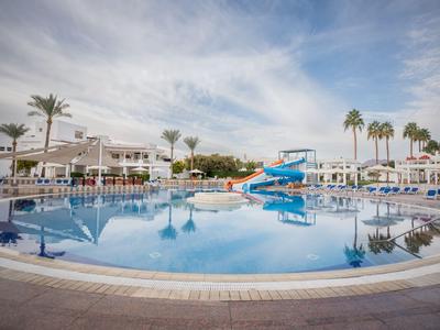 Großer Außenpool mit Wasserrutschen und Palmen in einer Hotelanlage bei bewölktem Himmel.