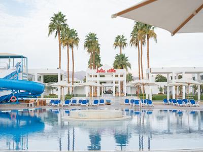 Großer Hotelpool mit Wasserrutsche, Liegestühlen und Palmen unter blauem Himmel.