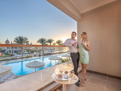 Paar genießt Getränke auf Hotelbalkon mit Blick auf Pool und Palmen bei Sonnenuntergang.