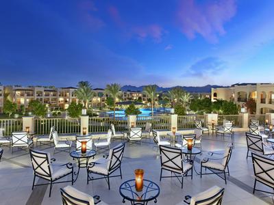 Offene Terrasse mit Tischen und Stühlen blickt auf Pool und Hotel bei Abenddämmerung.