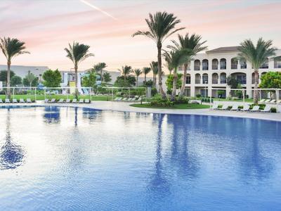 Großer Swimmingpool mit Liegestühlen und Palmen vor einem mehrstöckigen Hotelgebäude bei Sonnenuntergang.
