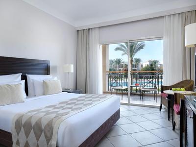 Helles Hotelzimmer mit großem Bett, Fliesenboden und Balkon mit Blick auf einen Pool.