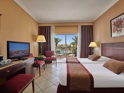 Hell eingerichtetes Hotelzimmer mit zwei Betten, Sitzbereich und Blick auf Terrasse und Palmen.