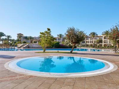 Runder Pool mit klarem Wasser vor Hotelgebäuden und Bäumen unter blauem Himmel.