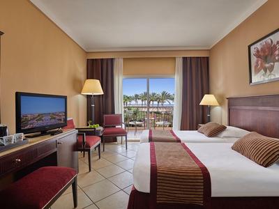 Hotelzimmer mit Doppelbett, Schreibtisch, Stühlen und Blick auf Balkon und Palmen.