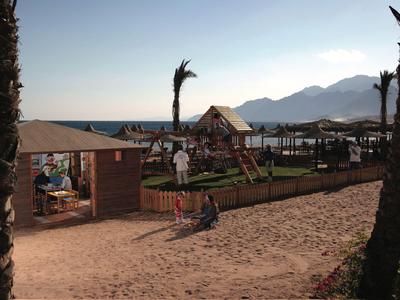 Strandbereich mit Sand, Palmen, Spielplatz und Meer im Hintergrund bei Sonnenuntergang.
