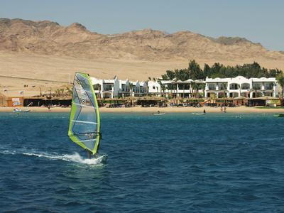 Ein Windsurfer fährt auf blauem Wasser vor einem weißen Hotel an einer Küste mit Bergen.