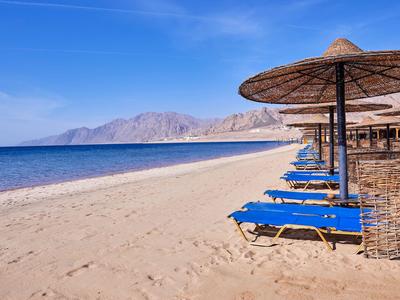 Leerer Sandstrand mit blauen Liegestühlen und Sonnenschirmen vor klarem blauem Himmel und Bergen.