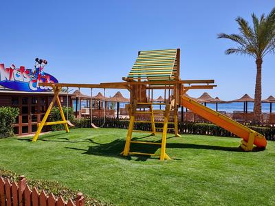 Spielplatz mit Rutsche und Schaukeln auf grünem Rasen neben Sonnenschirmen am Hotelstrand.