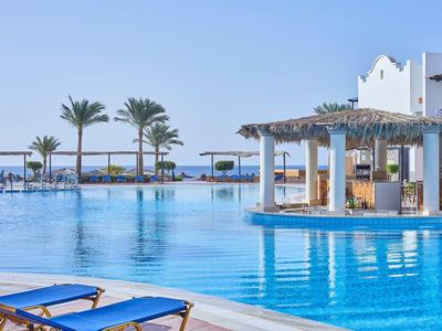 Luftiger Poolbereich mit Liegestühlen, Palmen und Bar in einem Hotel am Meer.