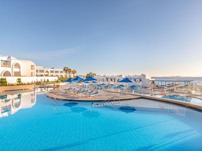 Großer blauer Swimmingpool vor weißen Hotelgebäuden bei klarem Himmel.