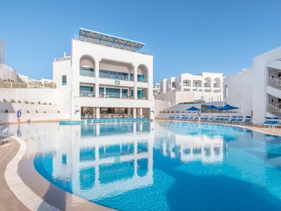 Modernes weißes Hotel mit großem beheiztem Pool und Sonnenliegen bei klarem Himmel.