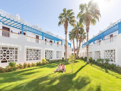 Zwei Personen entspannen auf einer grünen Rasenfläche zwischen weißen, zweistöckigen Hotelgebäuden unter blauem Himmel.