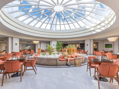 Helles Hotelrestaurant mit rundem Glasdach, bequemen Stühlen und Tischen in kreisförmiger Anordnung.