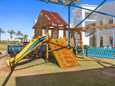 Bunter Kinderspielplatz mit Rutsche, Kletterwand und Schaukeln vor Hotelgebäude unter blauem Himmel.