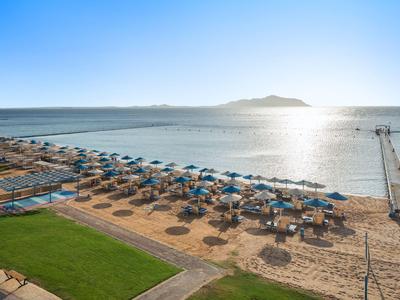 Strand mit Sonnenschirmen, Liegen und einem Pier bei ruhigem Meer und klarem Himmel.