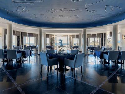 Elegante sala ristorante d'hotel con sedie blu, tavoli e grandi finestre con vista sul mare.