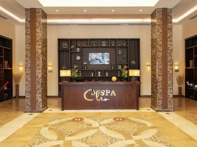 Hall d'hotel moderno con bancone reception, scaffali, illuminazione calda e tappeto decorativo.