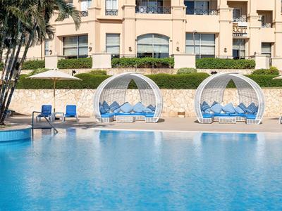 Grande piscina davanti a un hotel di lusso con letti lounge bianchi moderni e lettini blu.