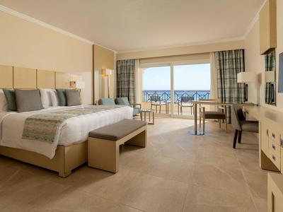 Camera d'hotel luminosa con grande letto, zona salotto e vista mare tramite ampie finestre.