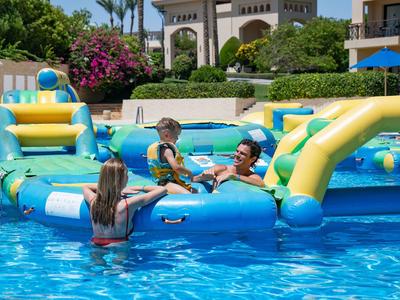 Famiglia che gioca su un gioco d'acqua gonfiabile in piscina in un hotel con ambiente tropicale.