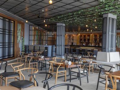 Modernes Hotelrestaurant mit Holz- und Metallmöbeln, großen Fenstern und grünen Pflanzen an der Decke.