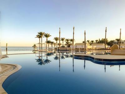 Poolbereich eines Hotels mit Palmen und Blick auf das Meer bei klarem Himmel.