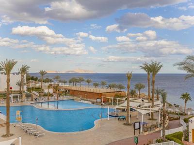 Hotelpool mit Sonnenliegen und Palmen, Blick auf das Meer und bewölkten Himmel.