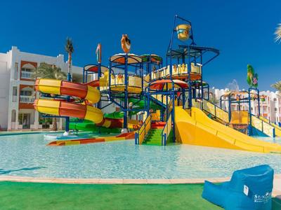 Wasserpark mit bunten Rutschen und Planschbecken vor einem Hotelgebäude und blauen Himmel.