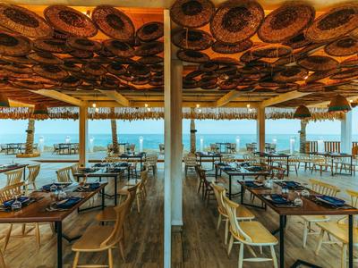 Helles Restaurant mit Holzstühlen und Tischen, offener Blick aufs Meer unter dekorativer Decke.
