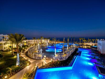 Hotelanlage mit beleuchtetem Pool und Palmen bei Nacht unter blauem Himmel.