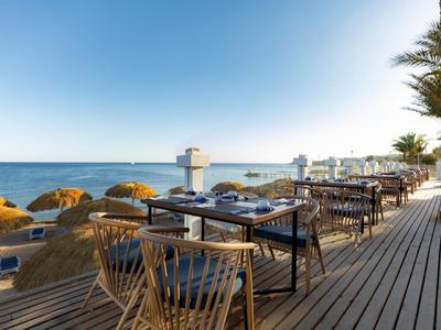 Terrasse mit Holzstühlen und Tischen mit Blick auf den Strand und das Meer bei klarem Himmel.