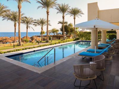Moderner Außenpool mit Liegestühlen und Blick auf Meer und Palmen in Hotelanlage.