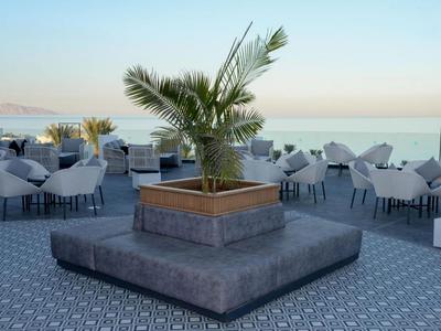 Terrasse mit Blick auf das Meer, Tische und Stühle mit weißen Tüchern, zentrale Palme in Blumenkasten.