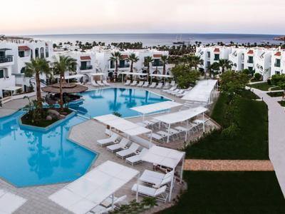 Großer Pool inmitten eines weißen Hotelkomplexes mit Liegestühlen und Grünflächen.